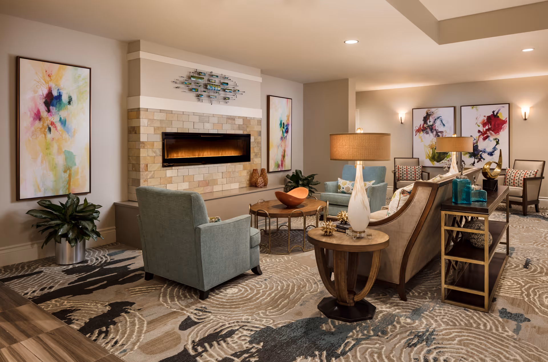 Well-lit sitting area with armchairs, a sofa, side tables and a modern fireplace, decorated with abstract art and lamps.