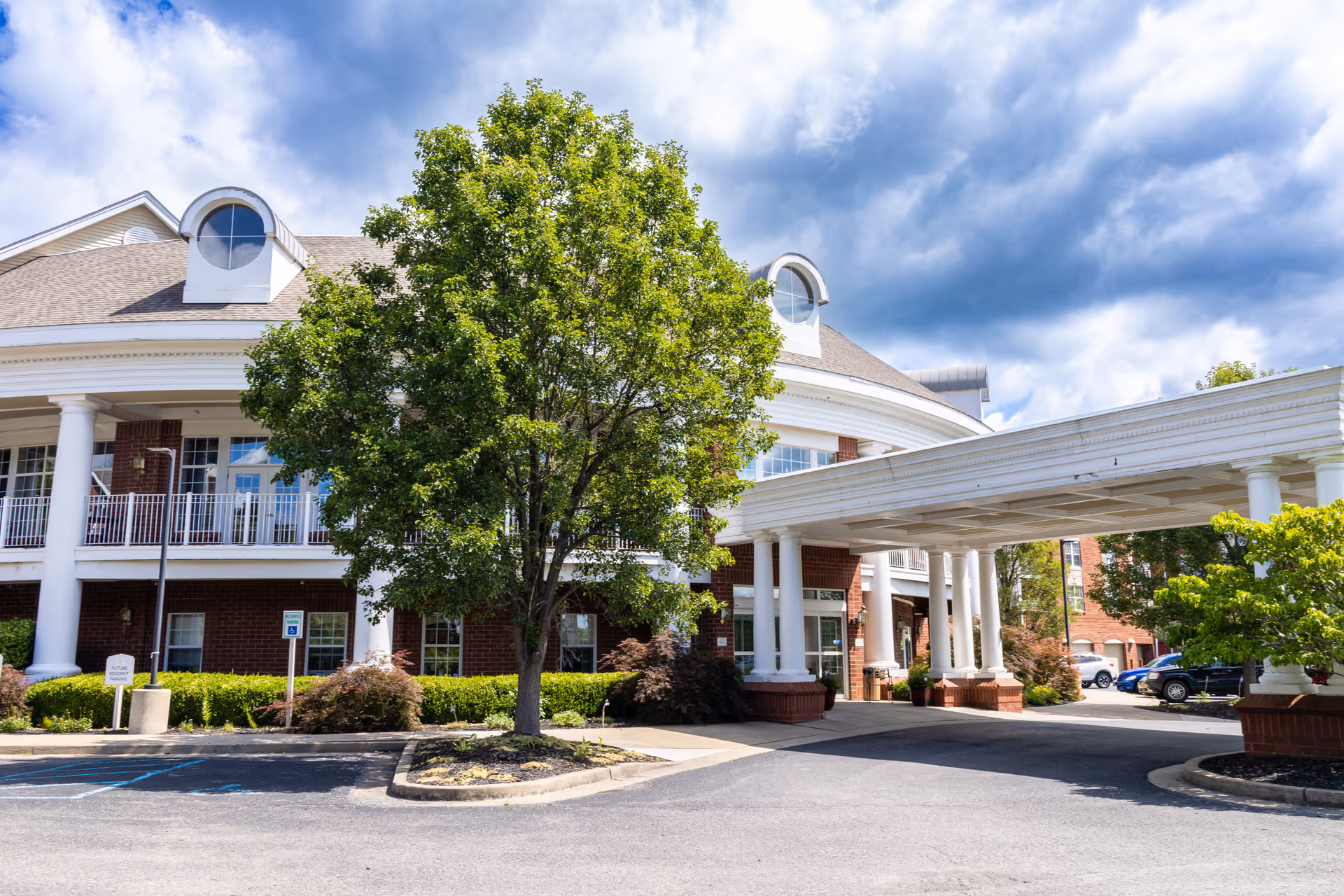 Exterior view of a senior living facility with a covered entrance supported by white columns, a large tree in front, and a brick building with white trim under a partly cloudy sky.