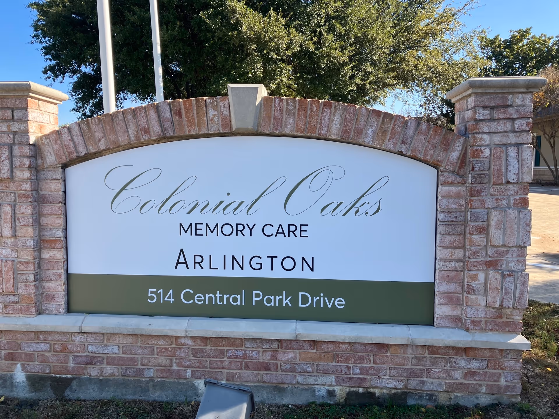 A brick sign for Colonial Oaks Memory Care Arlington located at 514 Central Park Drive, with trees and a clear blue sky in the background.