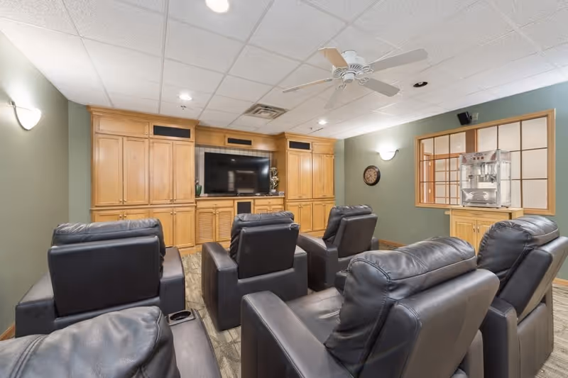 A cozy media room with six black leather recliners arranged in two rows facing a large flat-screen TV mounted on a wooden entertainment center. The room has green walls, a ceiling fan, wall sconces, a clock on the wall, and a popcorn machine on a wooden cabinet near a window.