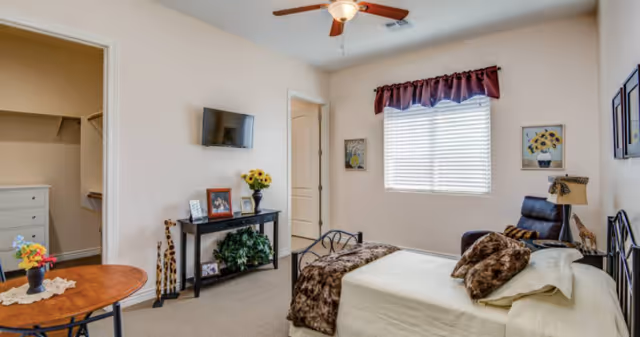 A bright and cozy bedroom with a bed covered in white and brown bedding, a small black metal table with a vase of flowers, a black console table with framed photos and a plant, a wall-mounted TV, a window with blinds and a maroon valance, and a ceiling fan. The room also has a walk-in closet visible through an open door.