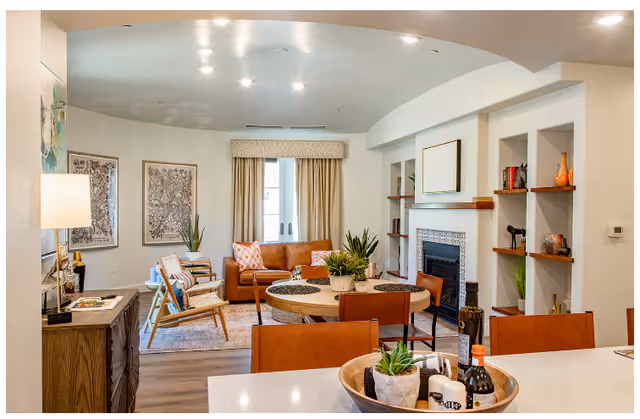 A cozy living room area with a brown leather sofa, two wooden chairs with cushions, a round wooden dining table with four chairs, and a fireplace with decorative tiles. The room has built-in shelves with various decorative items, a sideboard with a lamp, framed artwork on the walls, and a window with beige curtains. The foreground shows a kitchen counter with a tray holding plants and condiments.