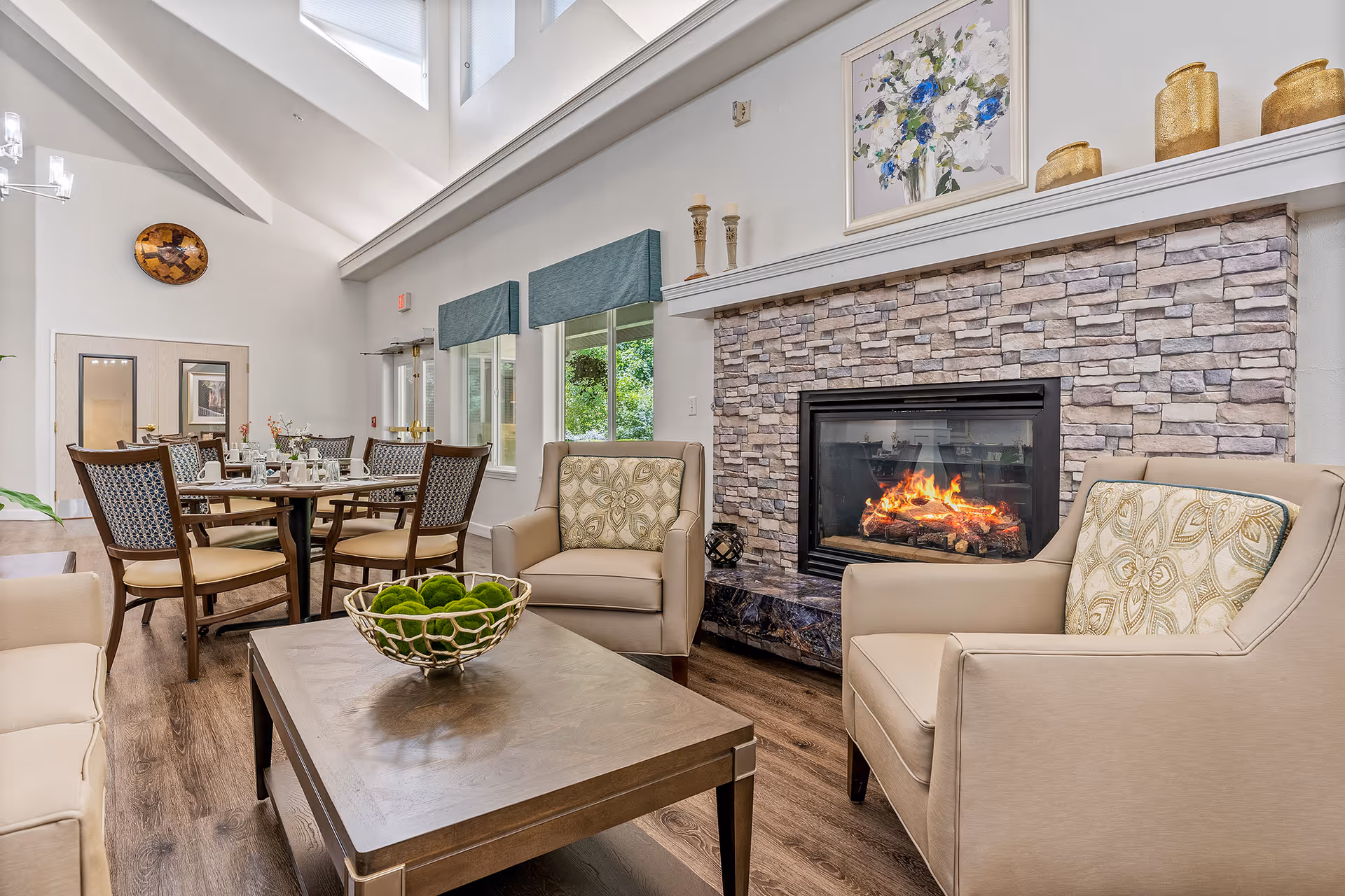 A cozy living room area in a senior living facility featuring a stone fireplace with a fire burning, two beige armchairs with patterned cushions, a wooden coffee table with a decorative bowl of green moss balls, and a dining table with chairs in the background. The room has high ceilings with windows near the roof, light-colored walls, and wooden flooring.