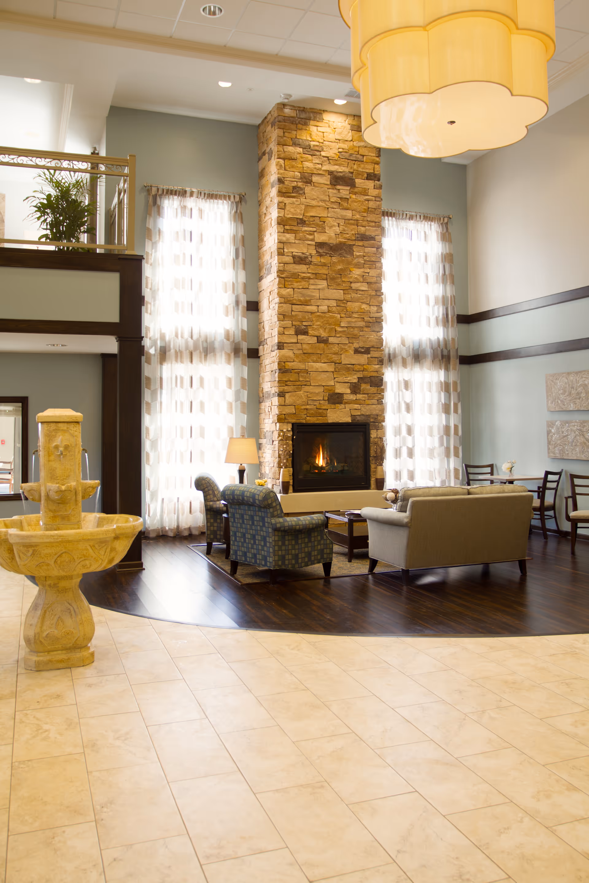 A bright lobby seating area with sofas and armchairs arranged around a tall stone fireplace, large windows, and a decorative fountain in the foreground.