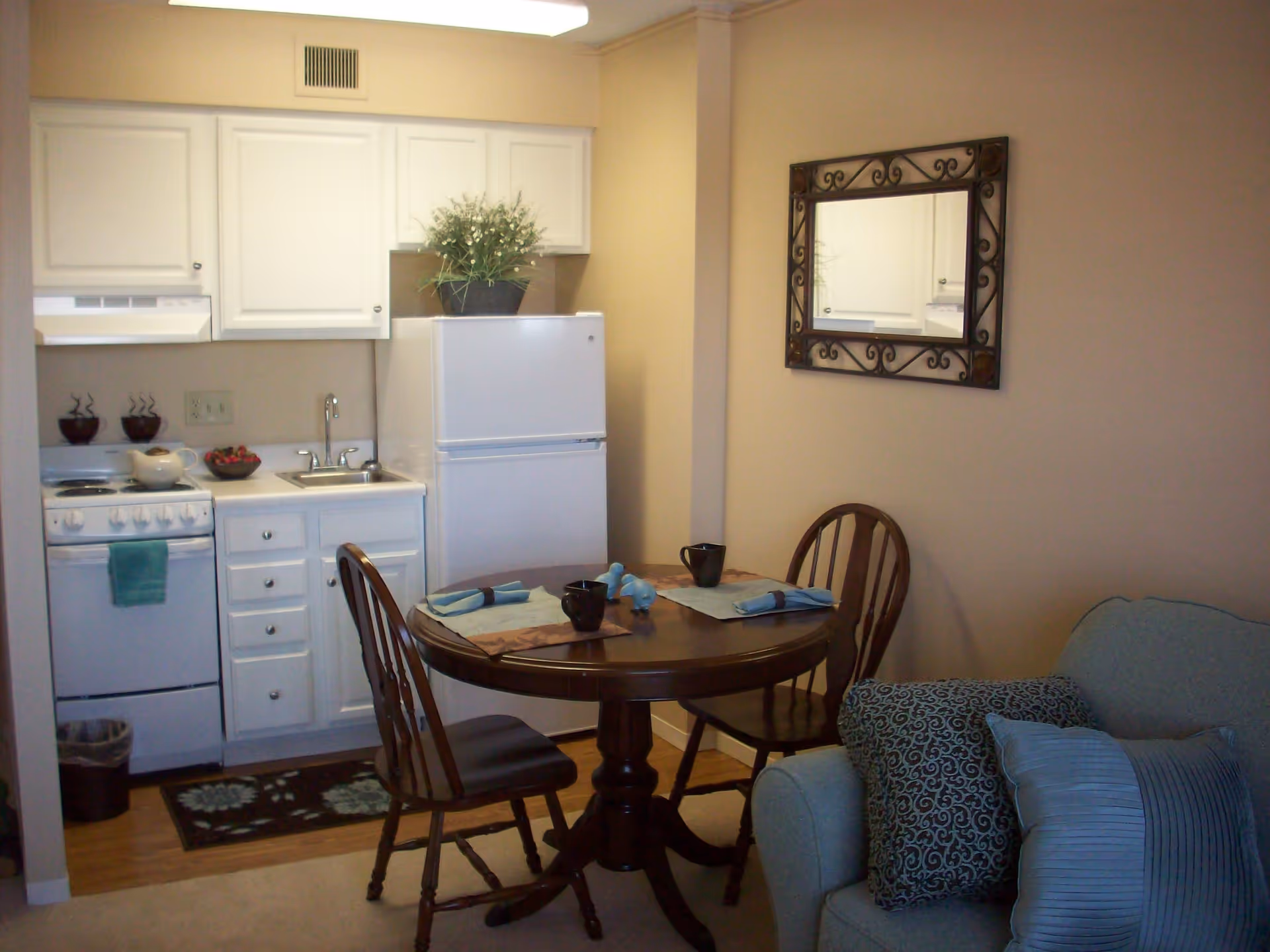 Small kitchen area with white cabinets, a white refrigerator, stove, and sink. A round wooden dining table with two chairs is set with placemats, napkins, and mugs. A decorative mirror hangs on the wall above a light blue couch with patterned pillows.