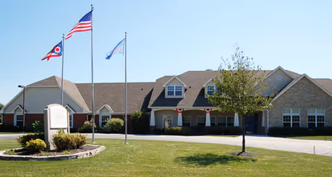 Exterior view of a single-story senior living facility building with a well-maintained lawn, three flagpoles displaying the American flag, Ohio state flag, and another flag, and a small tree in front of the building.