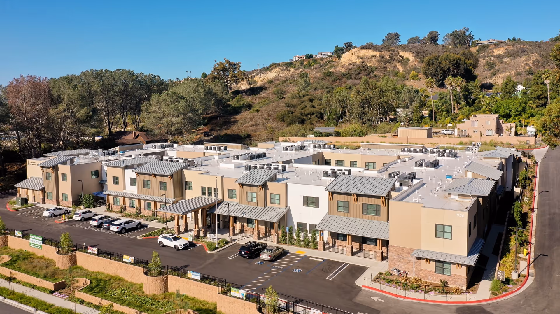 Aerial view of Westmont of Encinitas, a modern two-story senior living facility surrounded by trees and hills, with a parking lot in front and clear blue sky above.