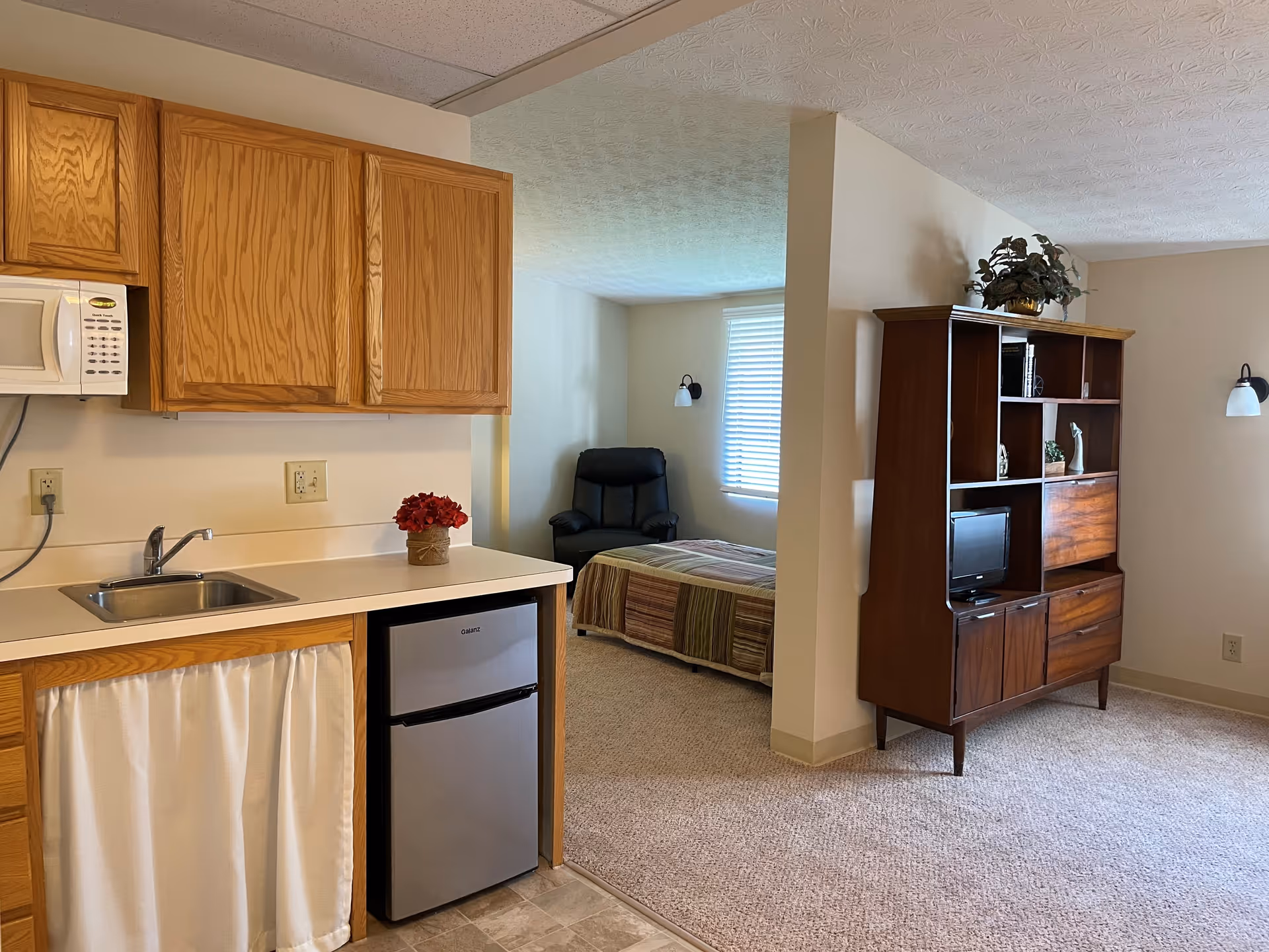 Interior view of a senior living facility room showing a small kitchenette with wooden cabinets, a microwave, a sink, and a mini refrigerator. Adjacent to the kitchenette is a bedroom area with a single bed covered with a striped blanket, a black armchair, and a window with blinds. A wooden shelving unit with decorative items and a small TV is positioned against the wall. The room has beige walls and carpeted flooring.