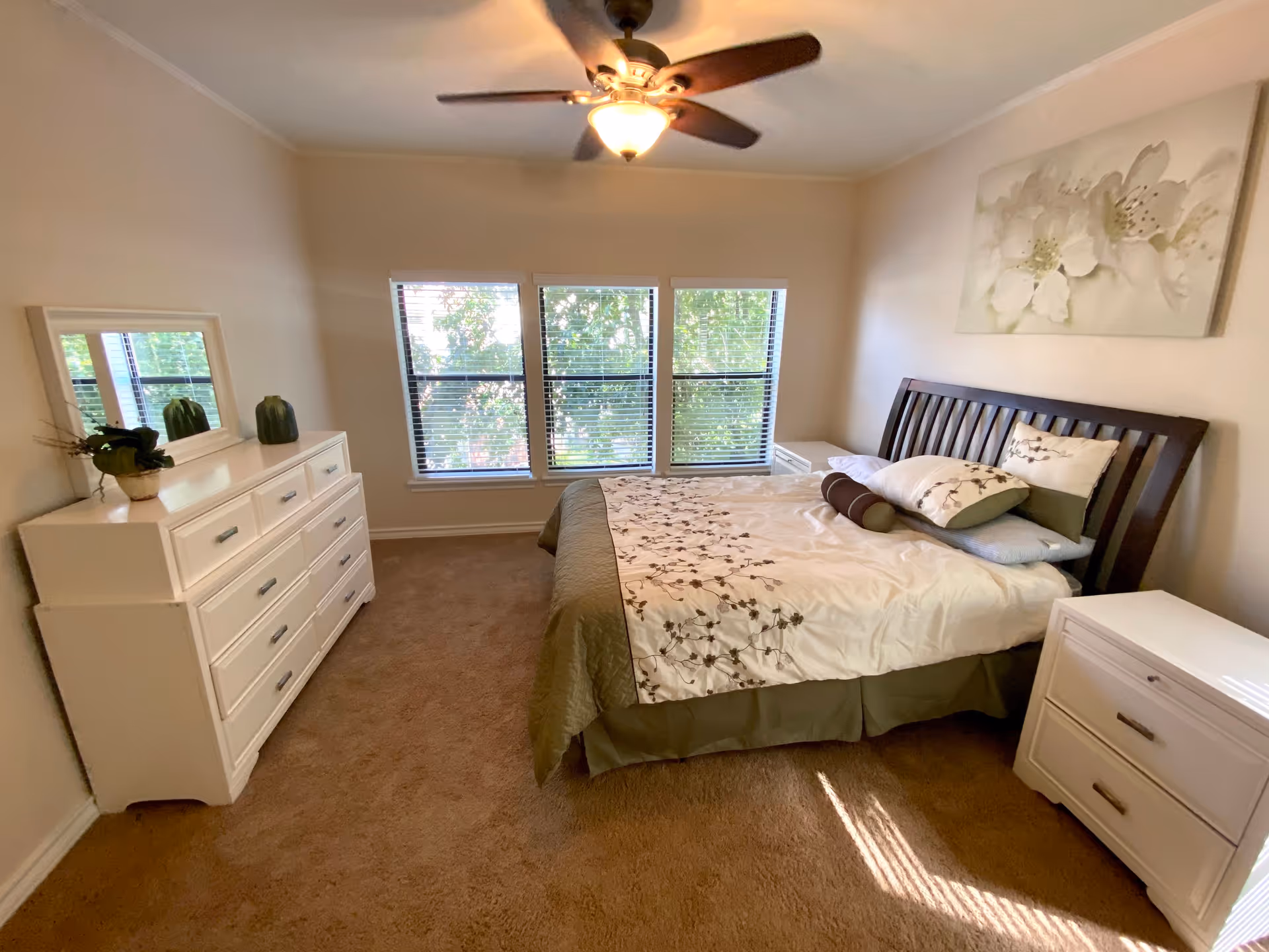 Bedroom with a wooden bed, white dressers, a ceiling fan, and three windows with blinds overlooking trees.
