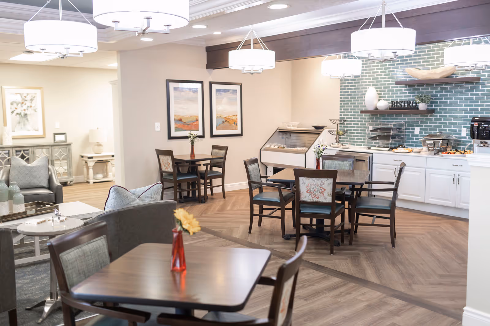 A bright and inviting dining area in a senior living facility with wooden tables and chairs, each table decorated with a small vase of flowers. The room features modern pendant lights, a green tiled backsplash behind a buffet counter with food trays, and framed landscape artwork on the walls. Adjacent to the dining area is a cozy sitting area with armchairs and decorative pillows.