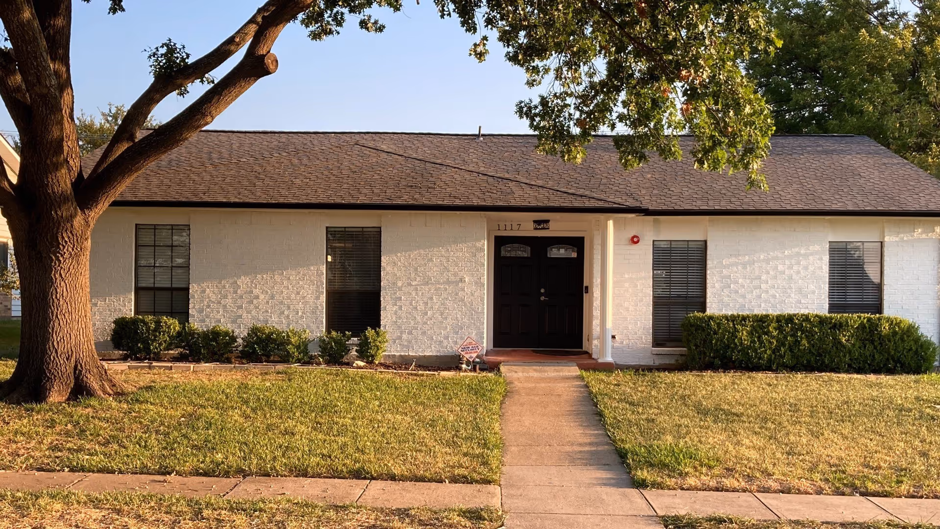Front exterior view of a single-story residential building with white brick walls, a dark shingled roof, and a black double front door. There are three windows with blinds, green bushes along the front, a large tree on the left side, and a concrete walkway leading to the entrance.