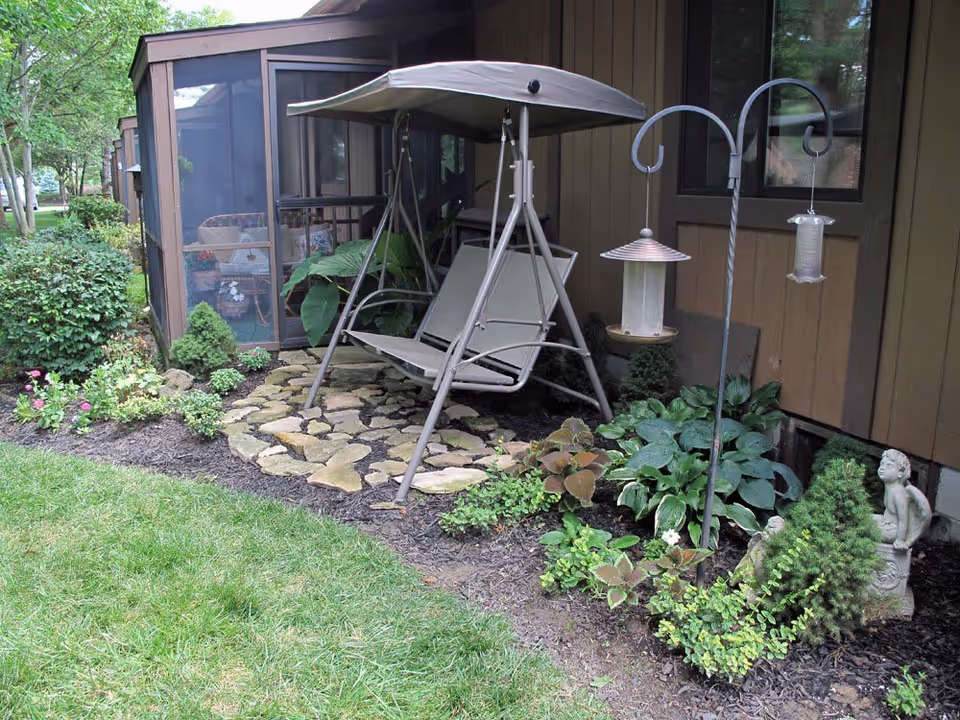 A garden area next to a building with a stone-paved section featuring a beige metal swing with a canopy. Surrounding the swing are various green plants and shrubs, a bird feeder stand with two feeders, and a small statue of a seated child. The building has brown wooden siding and screened-in porch sections.