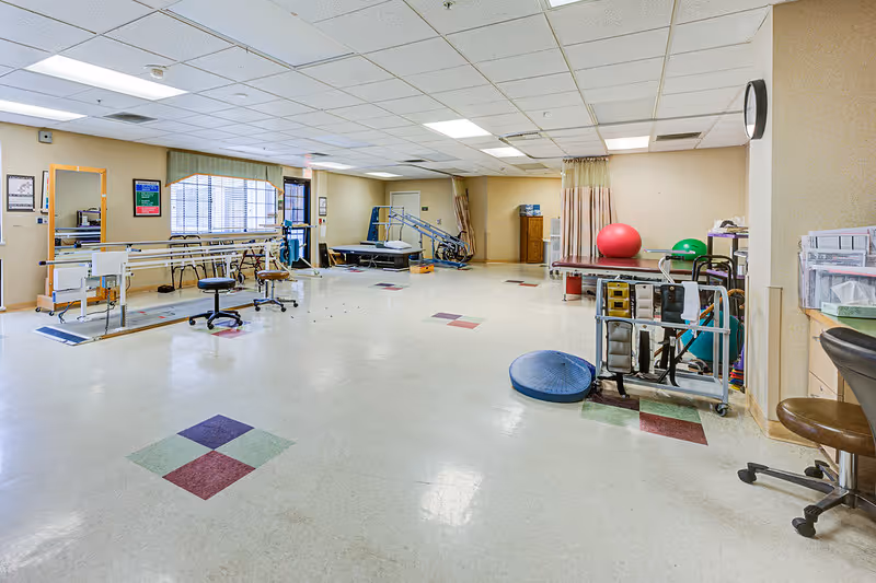 A spacious physical therapy or rehabilitation room with exercise equipment including parallel bars, therapy balls, a treatment table, chairs, and various therapy tools. The room has a tiled floor with colored square patterns and fluorescent ceiling lights.