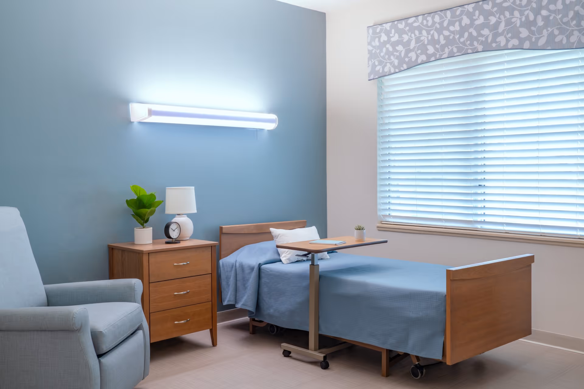A simple, clean bedroom in a senior living facility with a single wooden bed covered with a blue blanket, a small wooden nightstand with a plant, lamp, and clock, a light blue armchair, and a window with closed blinds and a floral valance.