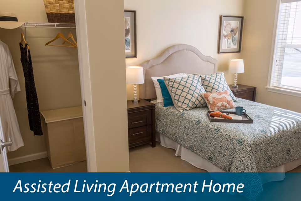 A furnished bedroom in an assisted living apartment with a made bed, nightstands, lamps, and an open closet.