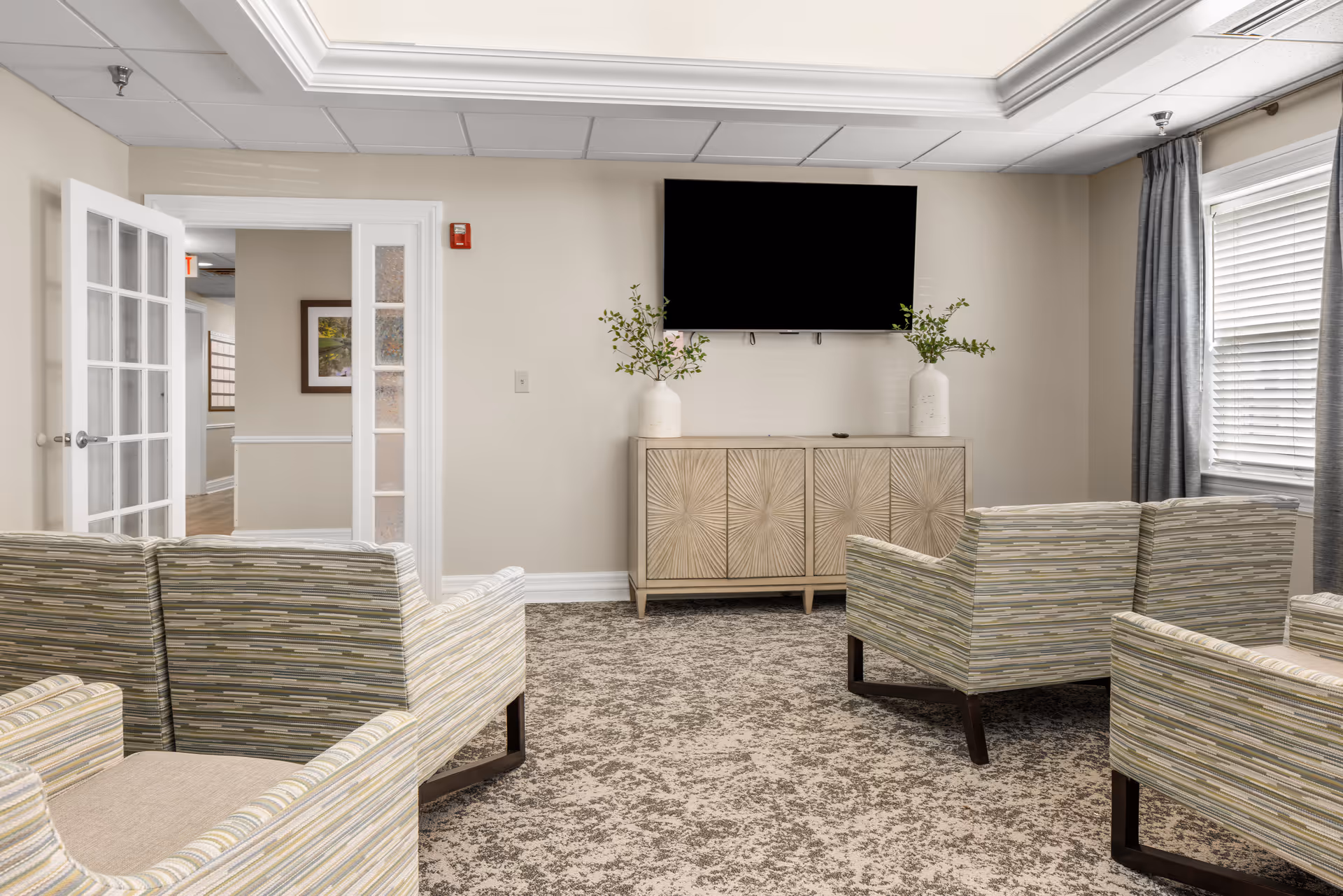 A cozy living room area with four striped armchairs arranged around a patterned carpet. A wooden cabinet with a sunburst design stands against the wall, topped with two white vases containing green leafy branches. Above the cabinet is a large flat-screen TV mounted on the wall. To the left, there is a white door with glass panels leading to a hallway. A window with gray curtains is on the right side of the room.