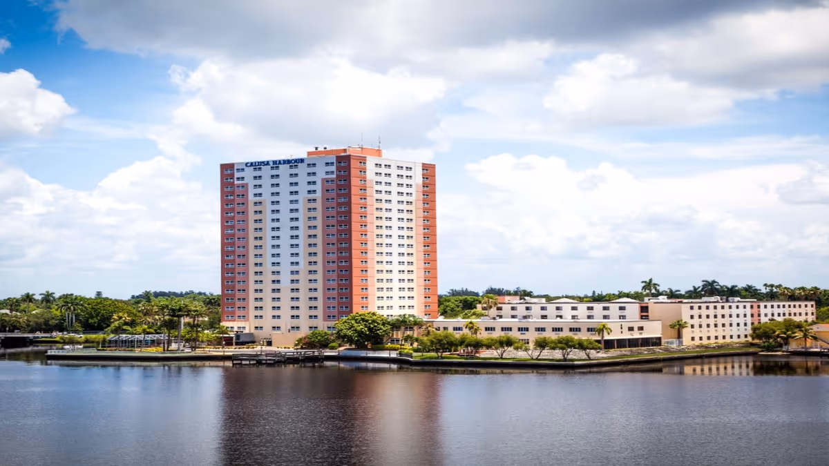 A tall multi-story building labeled Calusa Harbour situated beside a large body of water with a clear sky and scattered clouds in the background. The building is surrounded by greenery and smaller structures.