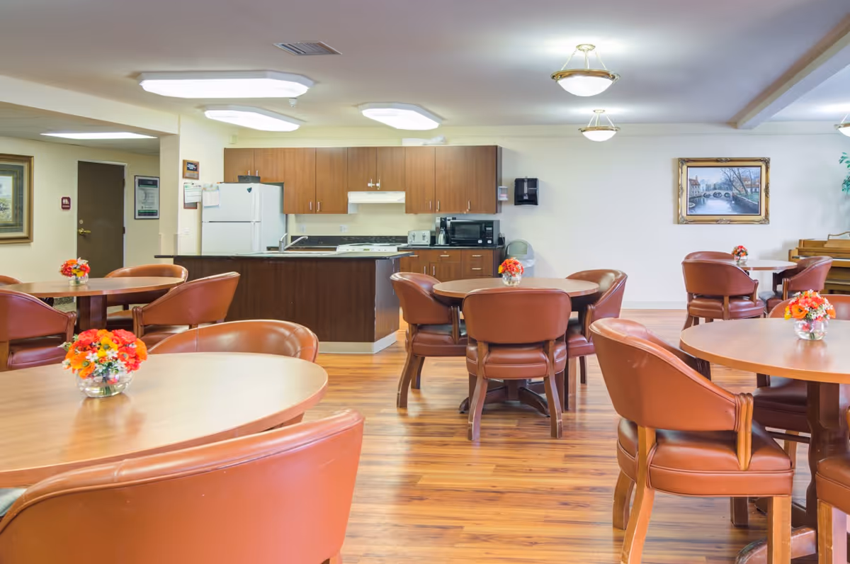 Community dining room with round wooden tables and brown leather chairs, small floral centerpieces, and a kitchenette in the background.
