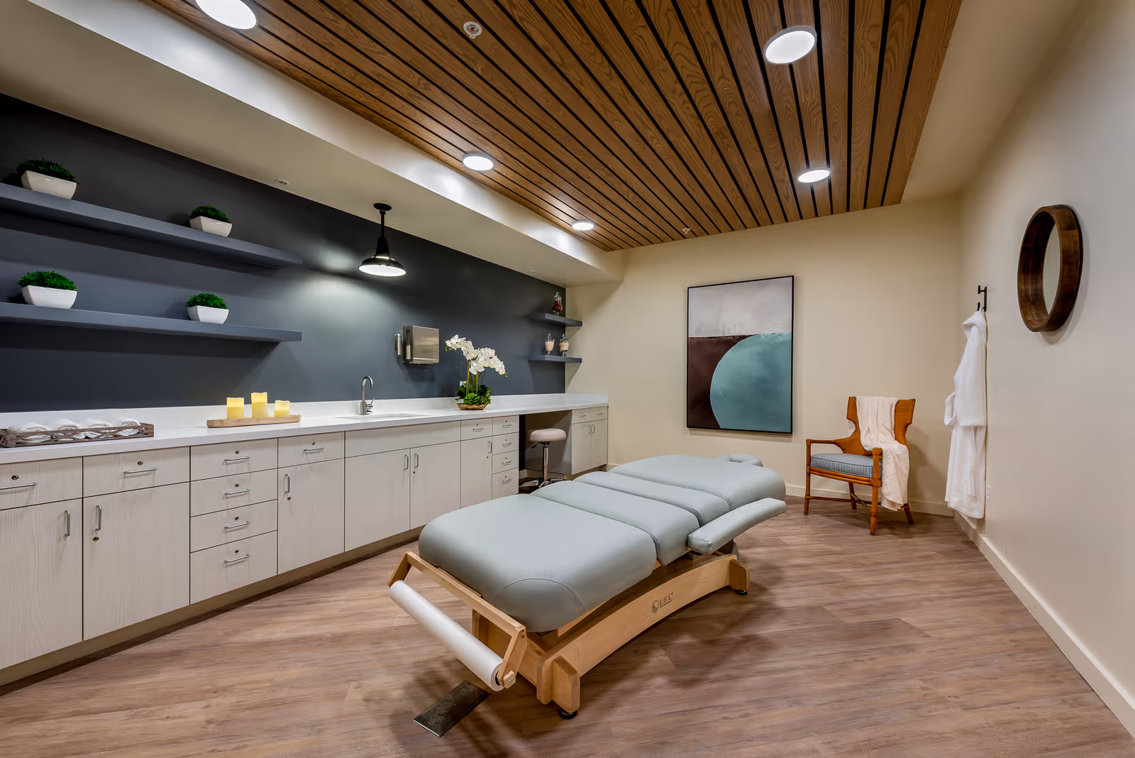 A modern therapy or massage room with a padded treatment table in the center, light wood flooring, a wooden slatted ceiling with recessed lighting, a long counter with cabinets and shelves holding small plants and candles, a chair with a white towel draped over it, a round mirror on the wall, and abstract artwork.