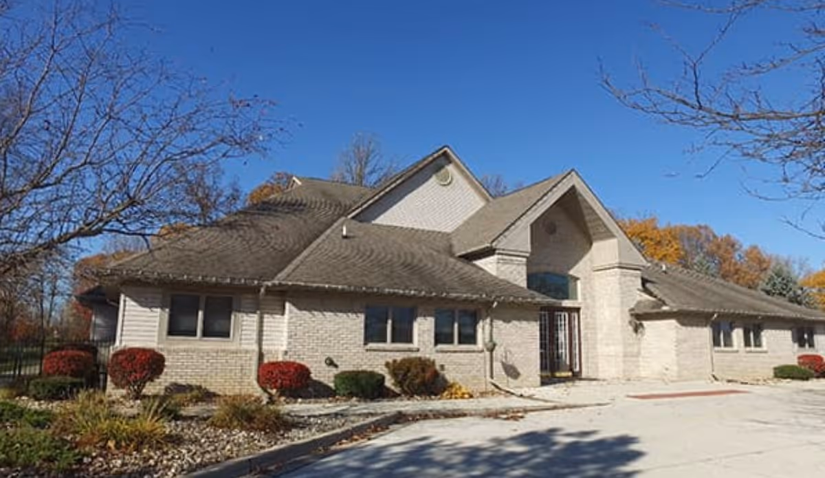 Exterior view of a single-story senior living facility building with a sloped roof, light-colored brick walls, and several windows. The building is surrounded by landscaped bushes and trees with autumn foliage under a clear blue sky.