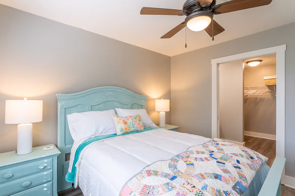 A bright bedroom with a light blue wooden bed frame and matching nightstands on each side. The bed is made with white bedding, a colorful quilt at the foot, and a floral decorative pillow. Two white lamps are on the nightstands, and a ceiling fan with a light is above. An open doorway reveals a walk-in closet with shelves and hanging space.