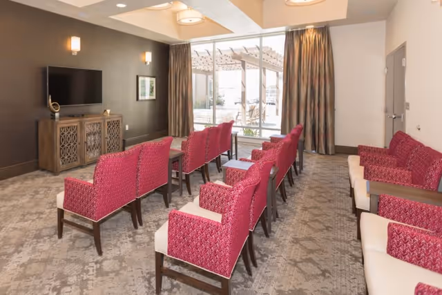 Interior common room with rows of pink upholstered chairs facing a wall-mounted TV and large window