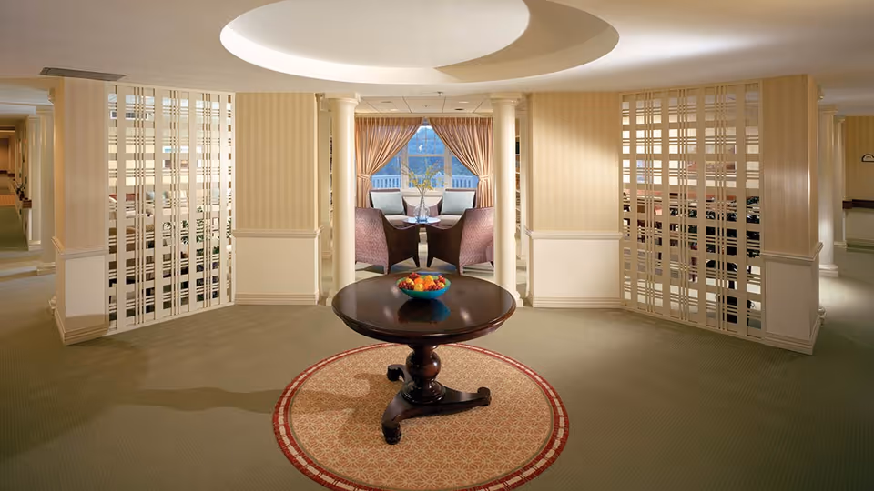 A spacious, well-lit interior area with beige walls and carpeted floors. In the center, there is a round wooden table with a bowl of fruit on top, placed on a circular patterned rug. Behind the table, two armchairs and a small table are positioned in front of a window with curtains. Decorative white lattice panels and columns frame the seating area.