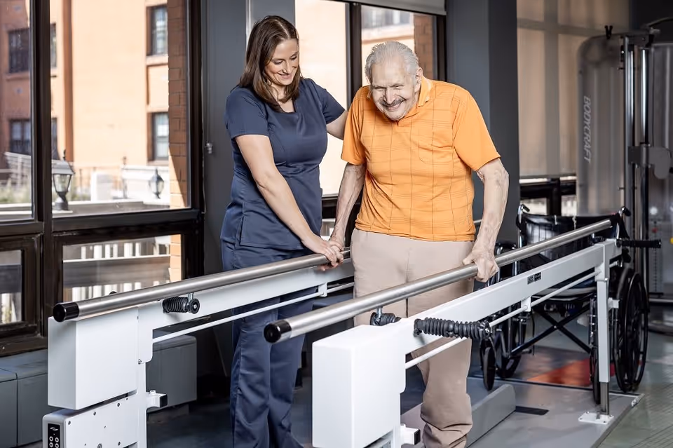 A female healthcare worker in navy scrubs assists an elderly man wearing an orange shirt and beige pants as he walks with support on parallel bars in a rehabilitation center with large windows and exercise equipment in the background.