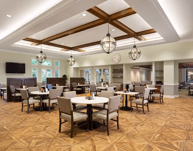A spacious dining room with multiple round tables and cushioned chairs arranged neatly. The room features a decorative coffered ceiling with wooden beams and hanging globe pendant lights. There is a wall clock above a built-in cabinet with glass doors, and a flat-screen TV mounted on the wall. The floor has a patterned wood design, and large windows allow natural light to enter the room.