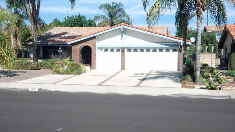 Single-story residential house front with a three-car garage, driveway, and palm trees.