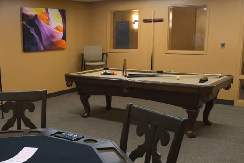 A game room with a pool table in the center, pool cues resting on the table, and a rack of billiard balls. There is a chair against the wall, two windows, and a colorful abstract painting on the wall. In the foreground, part of a card table with chairs is visible.