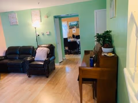 A cozy living room with light green walls, featuring a black leather sofa and armchair with a white blanket draped over the armchair. A floor lamp stands behind the armchair. To the right, there is a wooden piano with a plant and some items on top. The room has light wood flooring and an open doorway leading to another room with orange walls and a dining table with chairs.
