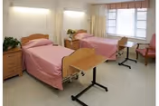 Two hospital-style beds with pink bedspreads in a nursing facility room with overbed tables, a nightstand, and a window.