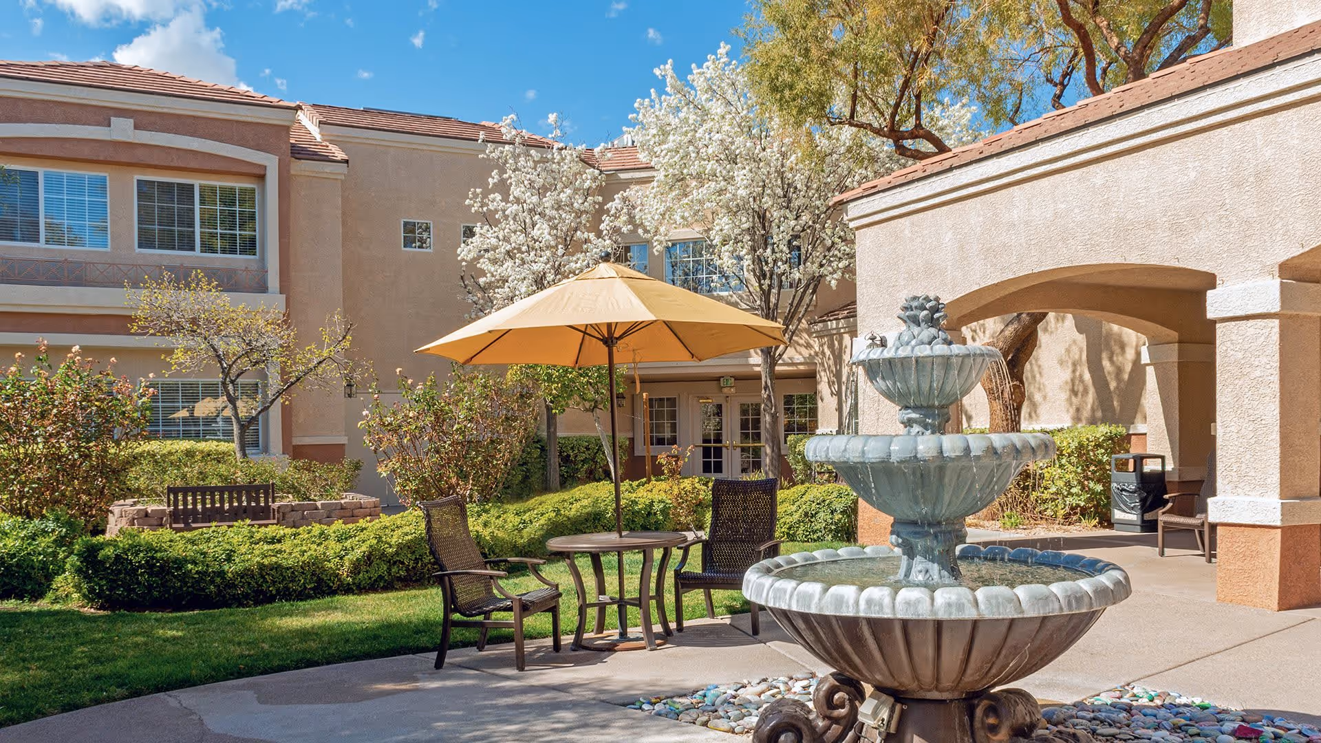 Outdoor patio area at a senior living facility with a three-tiered water fountain in the foreground, a round table with an umbrella, and two chairs. The background shows a beige building with windows, blooming trees, bushes, and a bench on a grassy area.