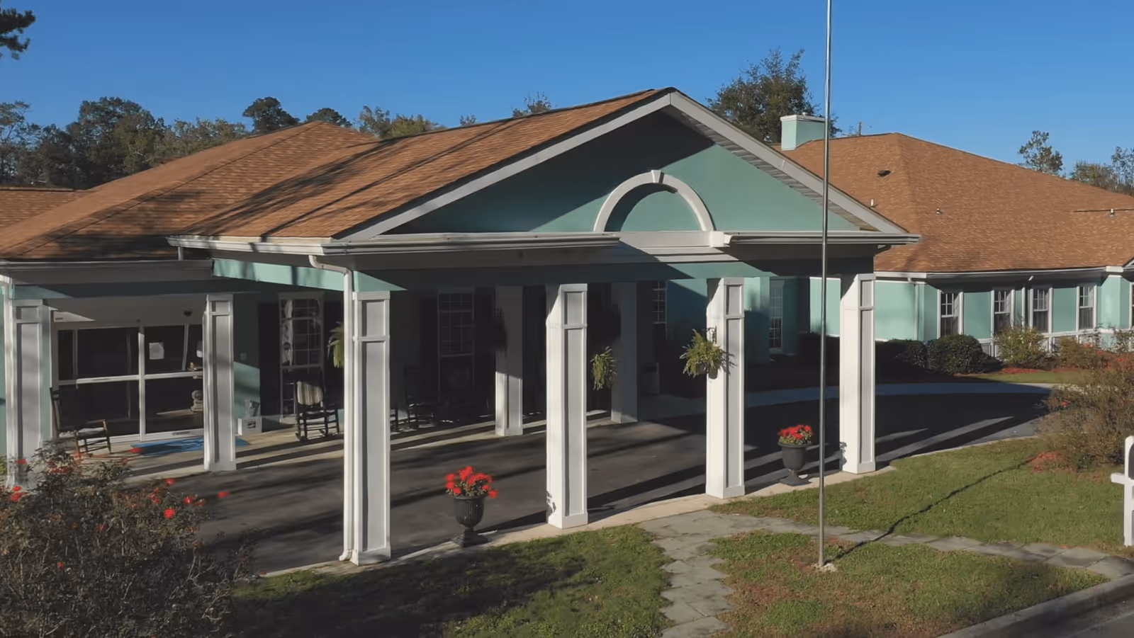 Exterior view of The Retreat at Summerville building entrance with a covered driveway supported by white columns, green walls, and a brown shingled roof. There are potted red flowers and hanging plants near the entrance, with a clear blue sky and trees in the background.