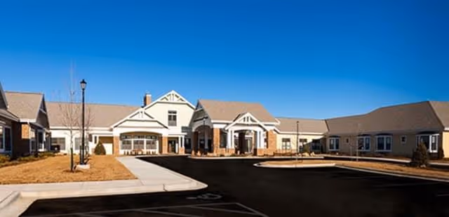 Exterior view of a single-story senior living facility building with beige siding and a gabled roof. The entrance features a covered portico with columns, and there is a paved driveway and parking area in front. The surrounding landscape includes dry grass and a few small trees.