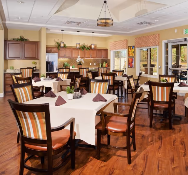A bright and spacious dining room with multiple tables covered in white tablecloths and set with folded brown napkins. Each table is surrounded by wooden chairs with striped cushions. The room features wooden flooring, a kitchen area with wooden cabinets and stainless steel appliances in the background, and large windows with patterned valances allowing natural light to fill the space. Decorative plants and framed artwork adorn the walls.