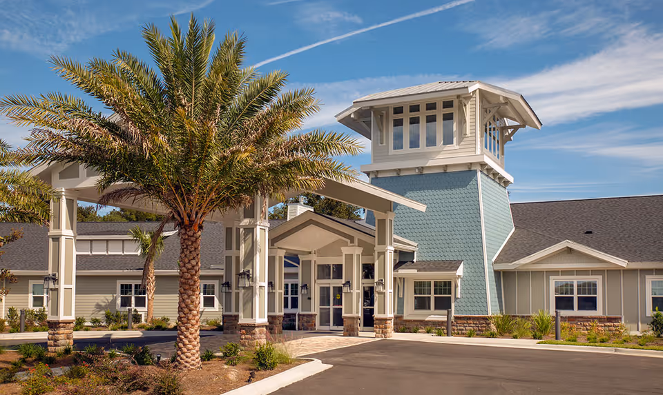 Exterior view of The Canopy at Harper Lake facility featuring a covered entrance with columns, a tall tower-like structure with multiple windows, palm trees, and landscaping under a blue sky with some clouds.