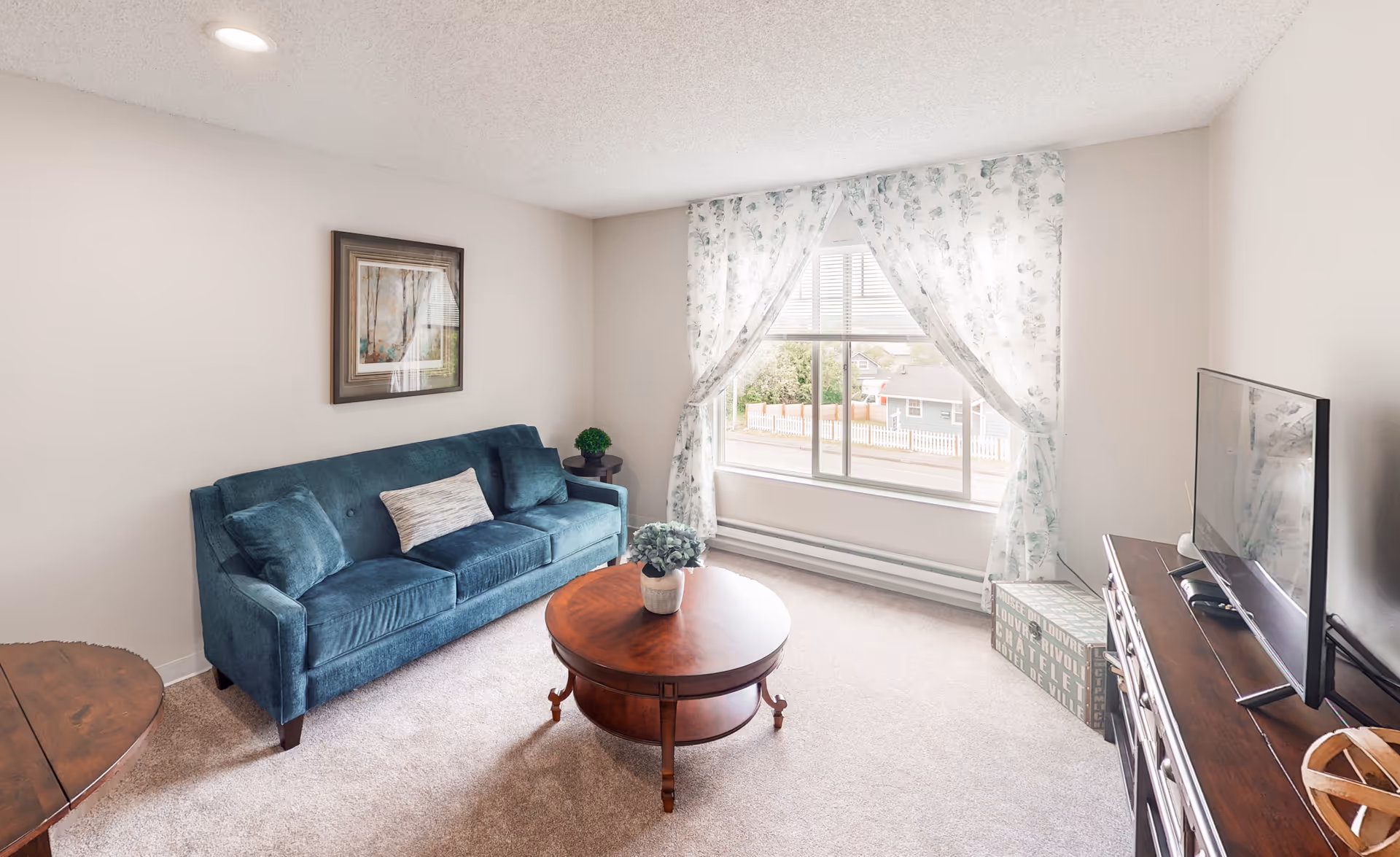 A bright living room with a teal sofa, a round wooden coffee table with a small plant, a large window with floral curtains, a wooden dresser with a flat-screen TV, and a framed painting on the wall.