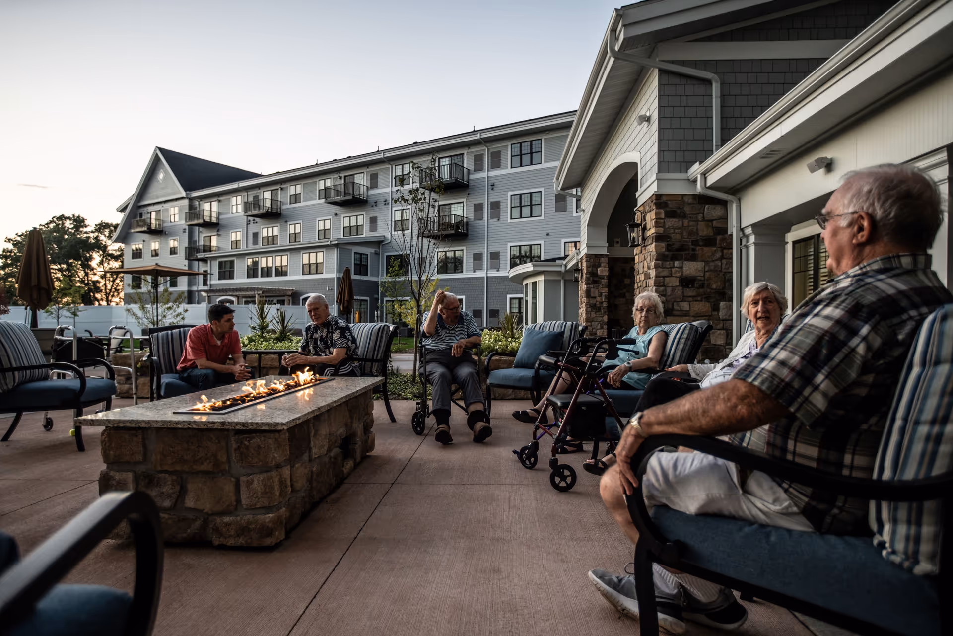 A group of elderly people sitting and chatting around a stone fire pit on an outdoor patio at dusk, with a multi-story residential building in the background.