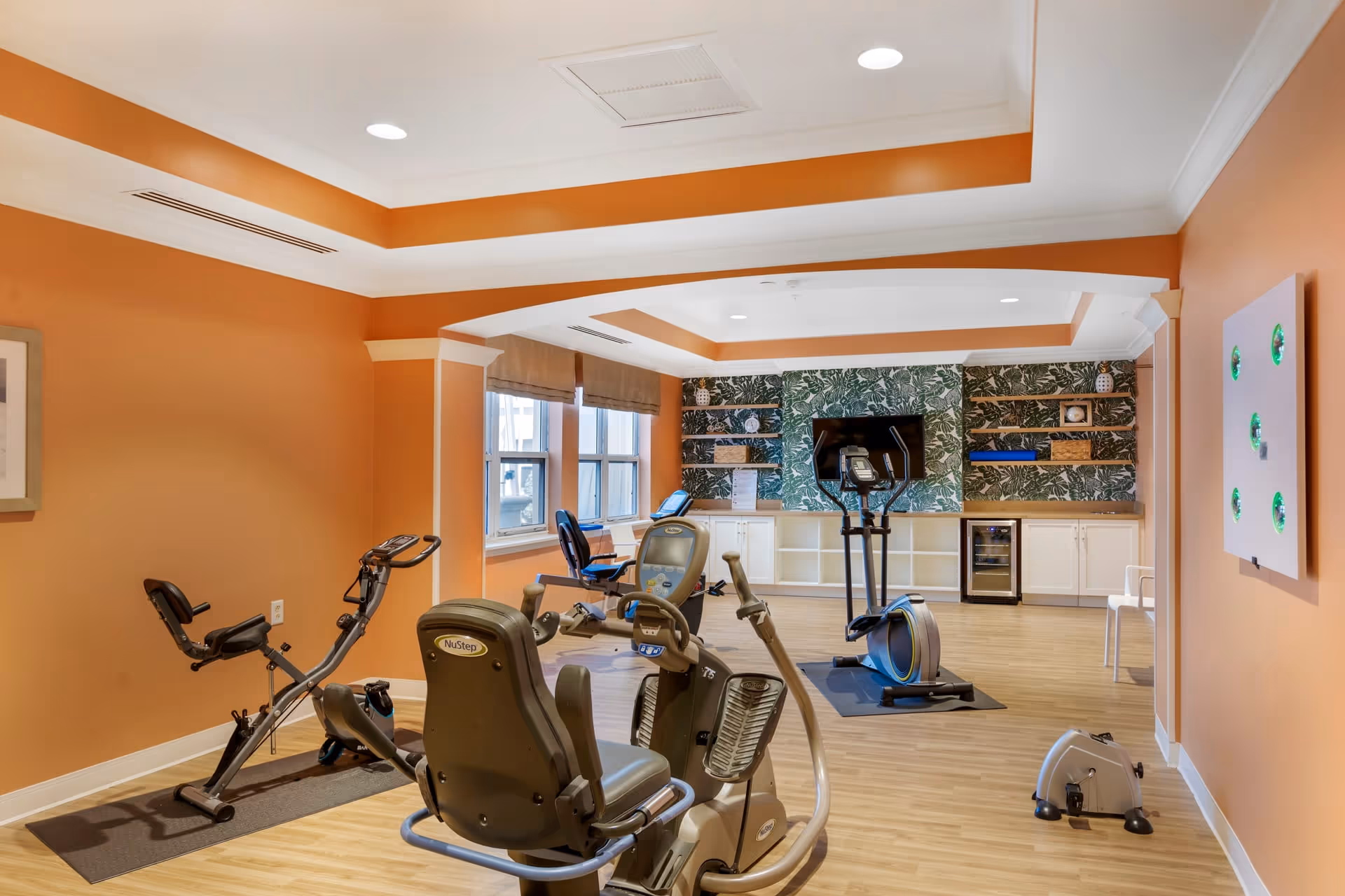 Exercise room with stationary bikes, an elliptical, and a TV mounted on a patterned accent wall in a senior living facility.