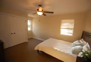 A bedroom with a bed covered in white bedding and several pillows, a ceiling fan with a light fixture, two windows with blinds, and a closed double-door closet.