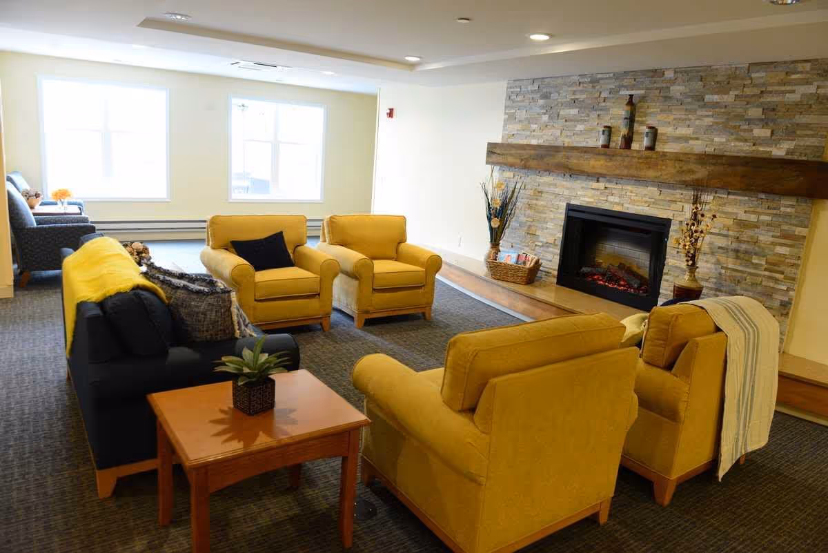 A cozy living room with four mustard yellow armchairs arranged around a wooden coffee table with a small potted plant. There is a black couch with a yellow throw blanket and pillows. The room features a stone fireplace with a wooden mantle decorated with vases and candles. Two large windows allow natural light to fill the space.
