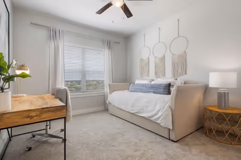 A bright bedroom with a large window covered by sheer curtains, a beige daybed with white bedding and decorative pillows, three macrame wall hangings above the bed, a wooden desk with a plant and lamp, a ceiling fan, and a round side table with a lamp.