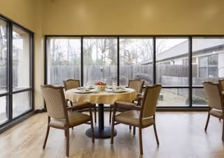 Dining area with a round table set for a meal and upholstered chairs in front of large floor-to-ceiling windows overlooking a fenced courtyard.