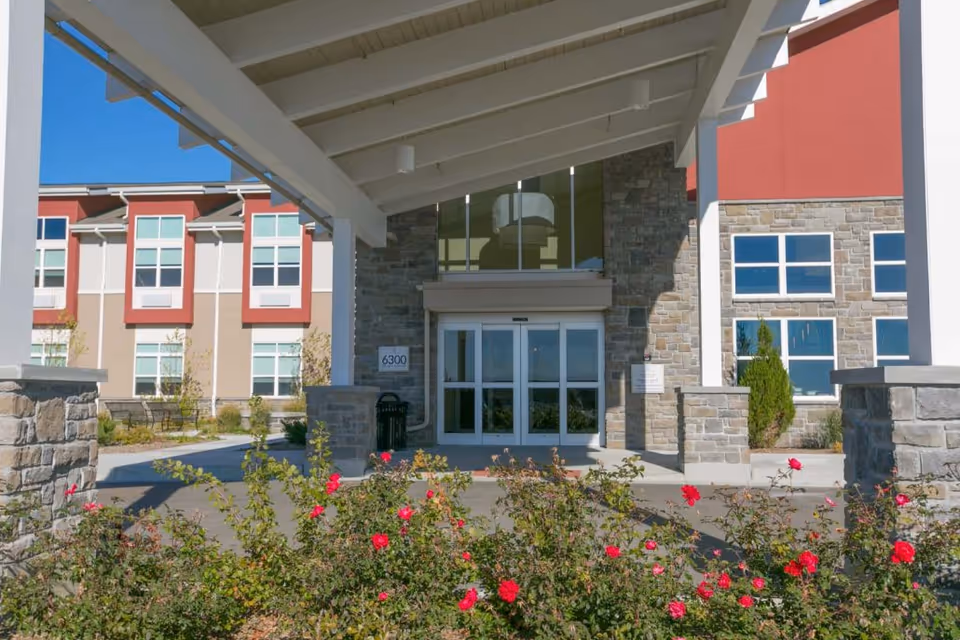 Entrance of a senior living facility with a covered driveway supported by white beams. The building has stone and beige walls with red accents around the windows. There are bushes with red flowers in the foreground and clear blue sky above.
