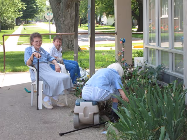 Three elderly women outside near a garden area. Two women are sitting on chairs on a sidewalk, one holding a cane and smiling, while the third woman is kneeling and tending to the plants in the garden bed next to a building with large windows.