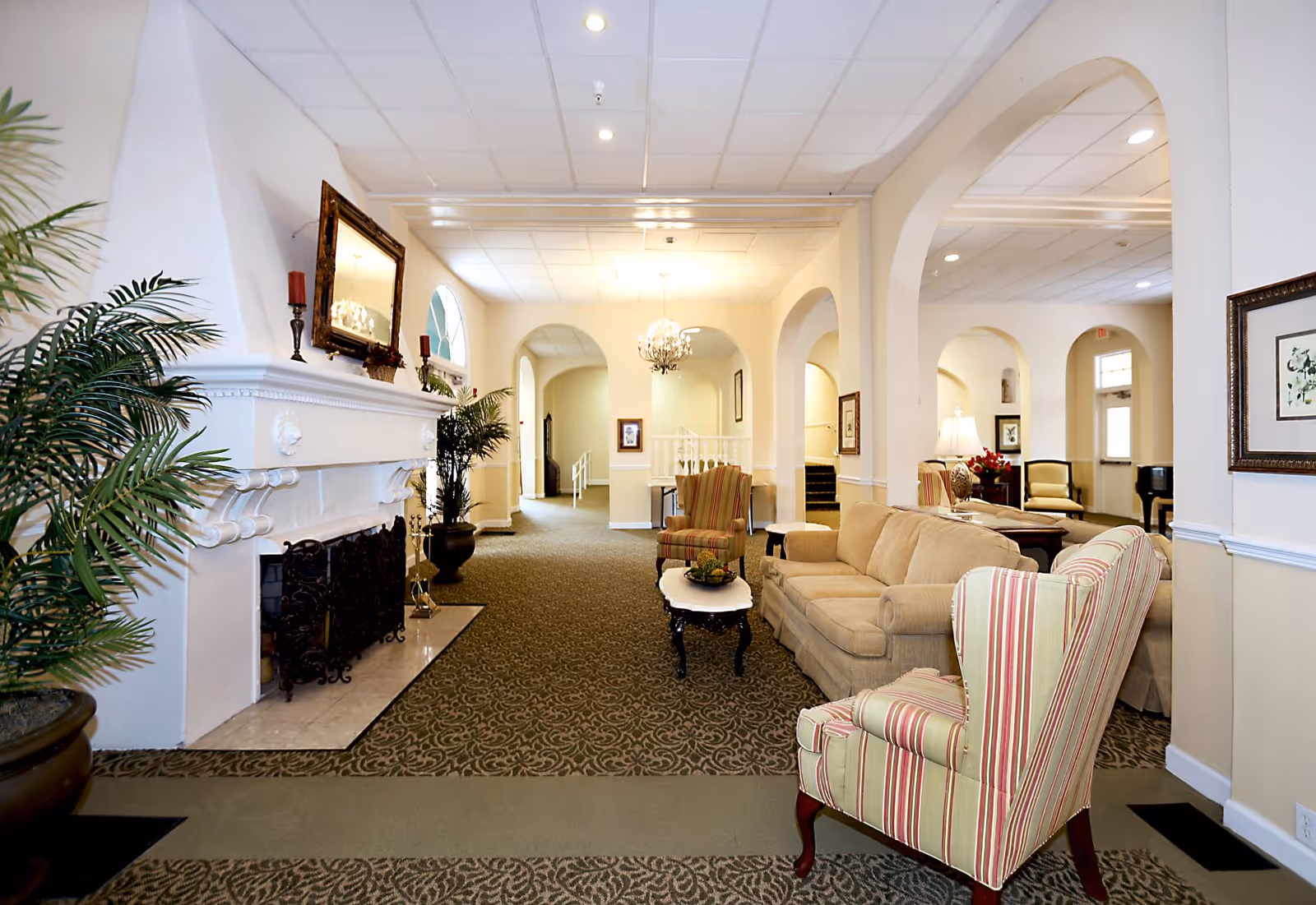 Bright, elegant senior living lounge with sofas, striped armchairs, a fireplace, and arched walkways.