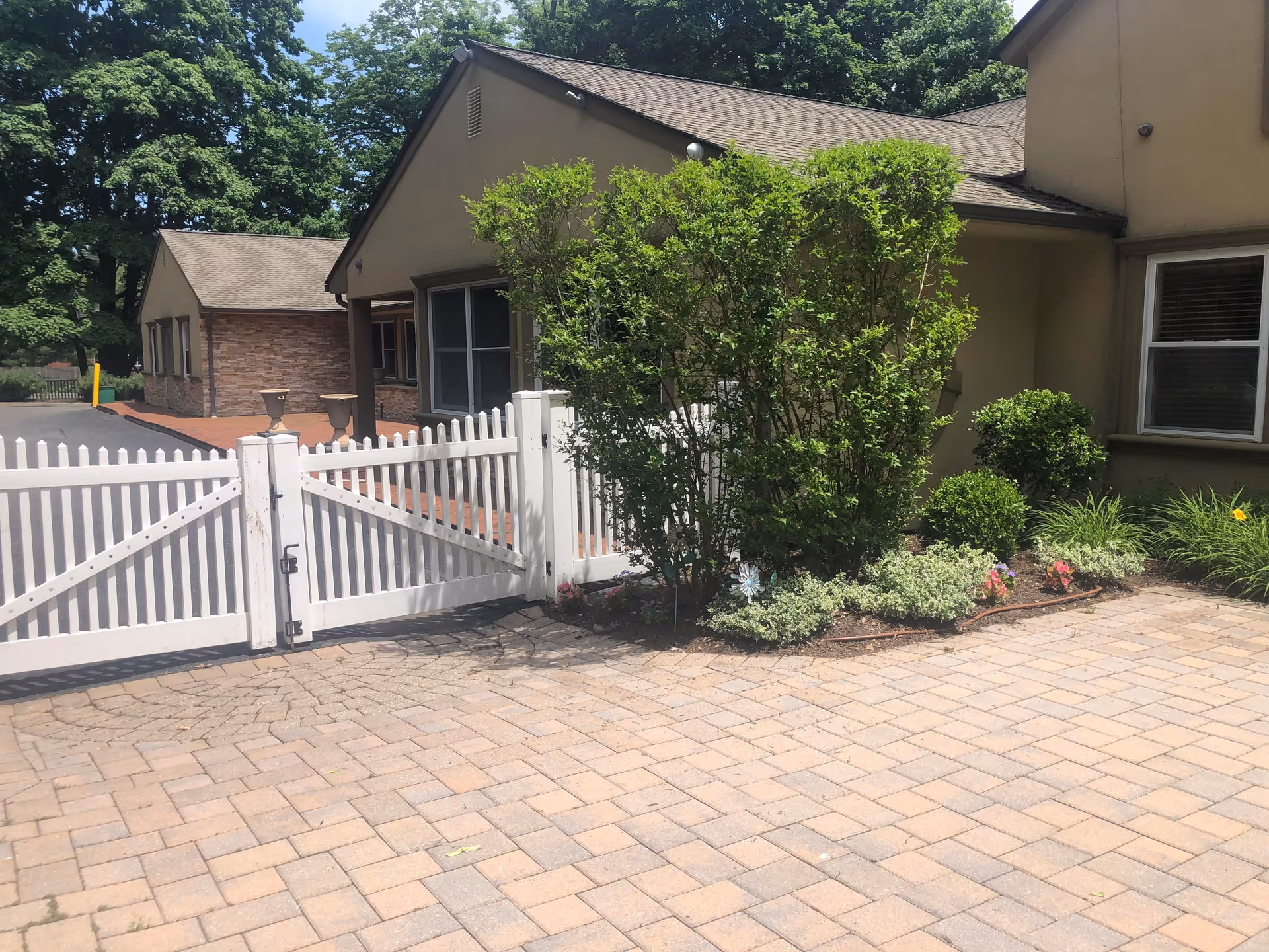 Exterior view of a building with beige walls and a brown roof, featuring a white picket fence gate and a paved driveway. There are green bushes and plants along the side of the building, with trees in the background under a clear sky.