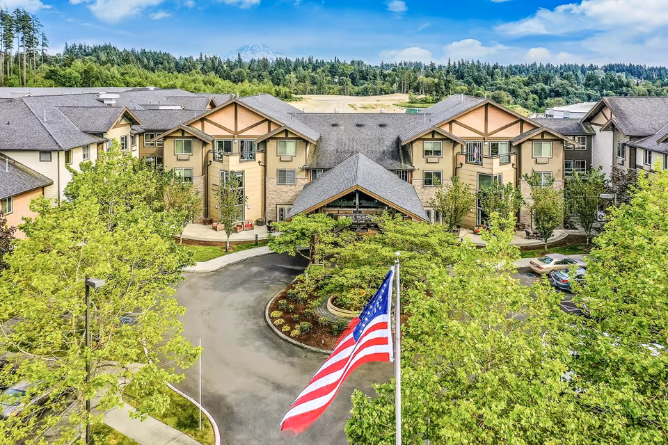 Exterior view of Meridian at Stone Creek senior living facility surrounded by green trees and landscaping, with an American flag prominently displayed in the foreground and a mountain visible in the background under a blue sky.